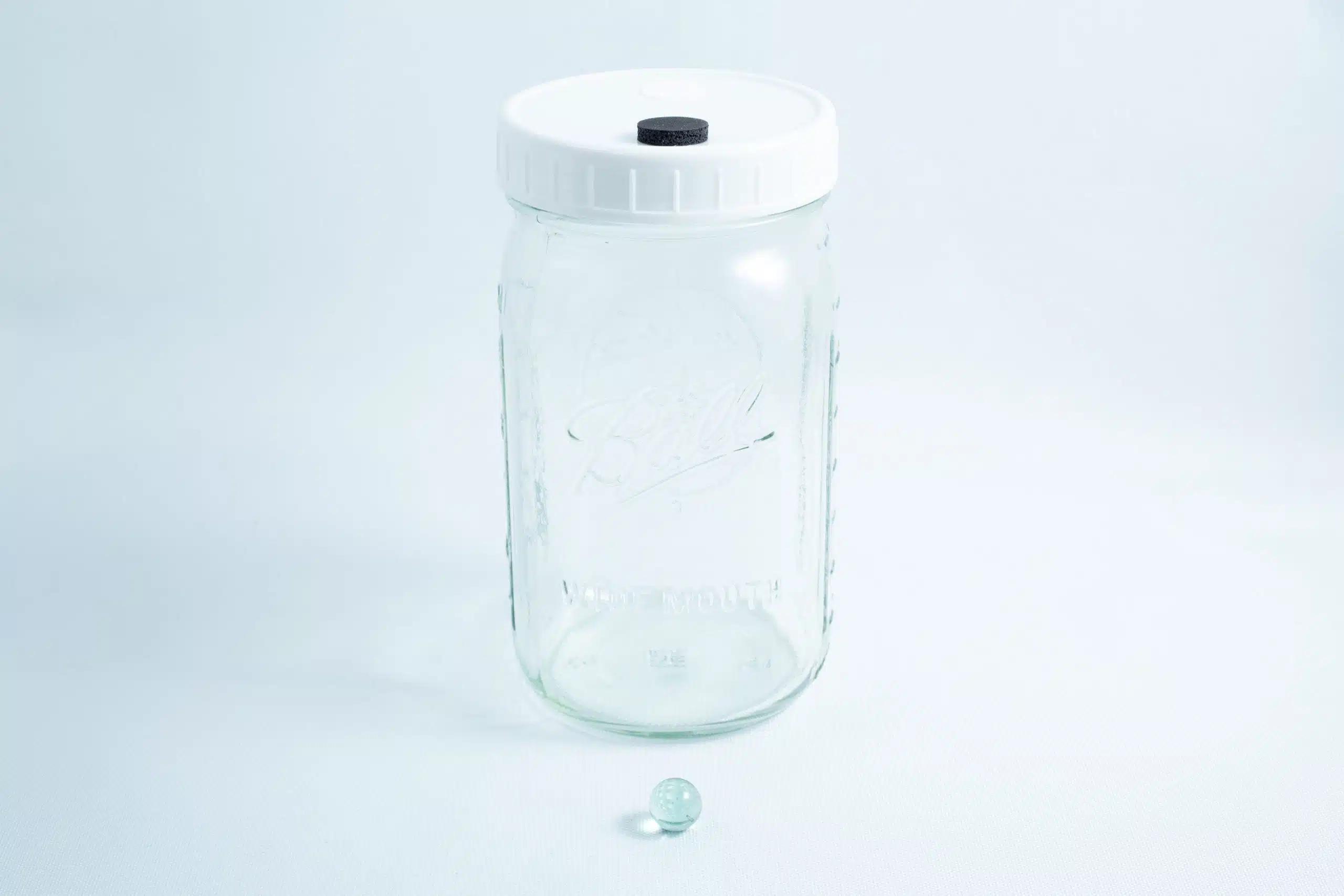A Mycology Ball Kilner jar with injection port and air vent, 1L genuine and autoclavable, sits next to a small glass marble on a white background.
