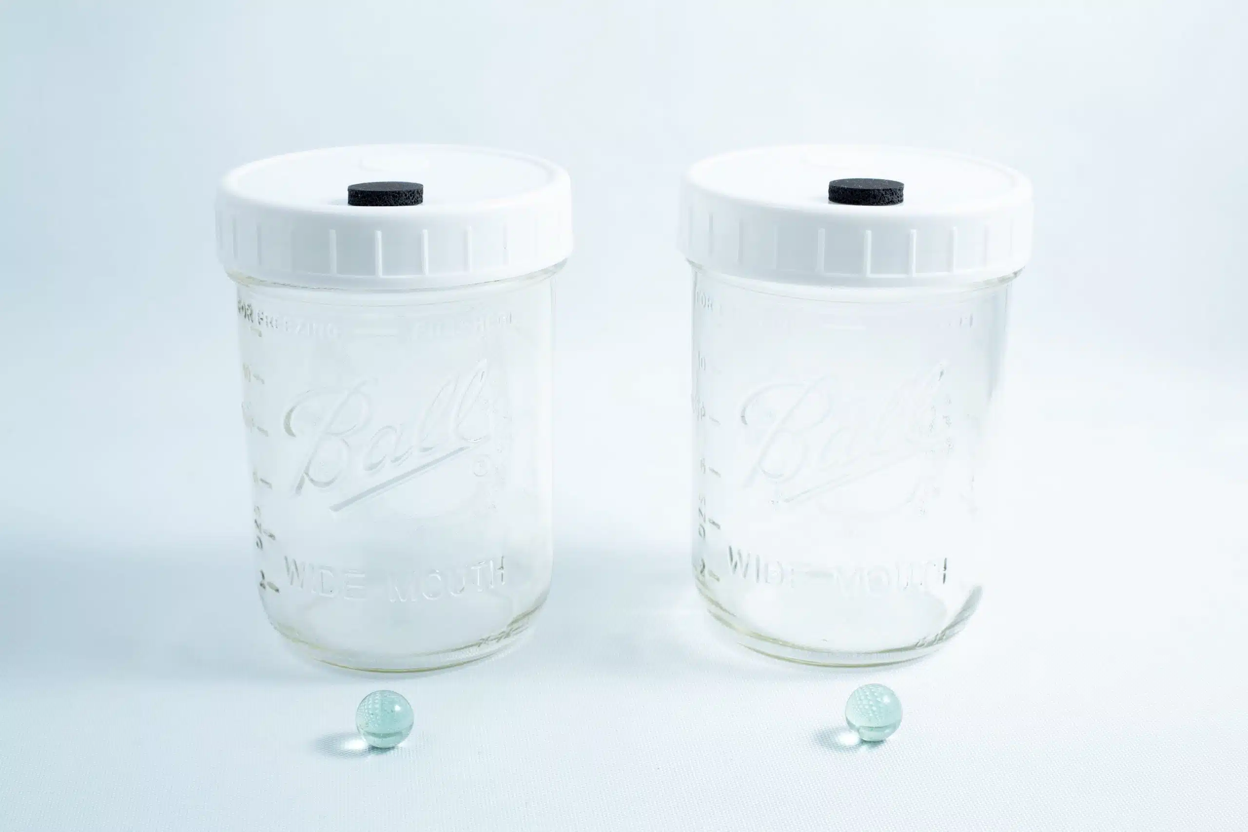 Two empty Mycology Ball Mason jars with injection ports and air vents, 1L genuine autoclavable, sit side by side, green marbles in front on a clean white background.