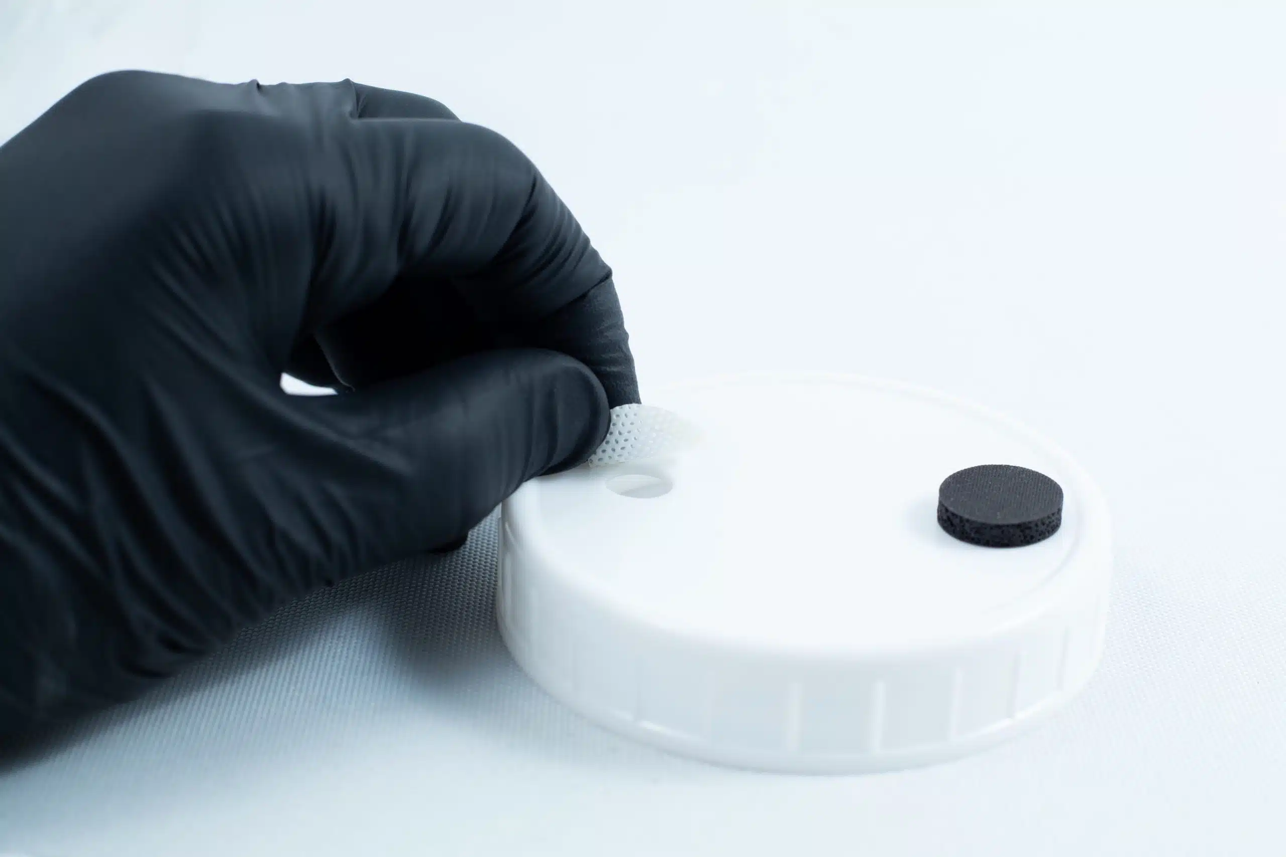 A gloved hand positions a mesh disc into a hole on a white lid beside a black disc, preparing the area for adhesion using Primer 94 multisurface for 3M glue 10ml.