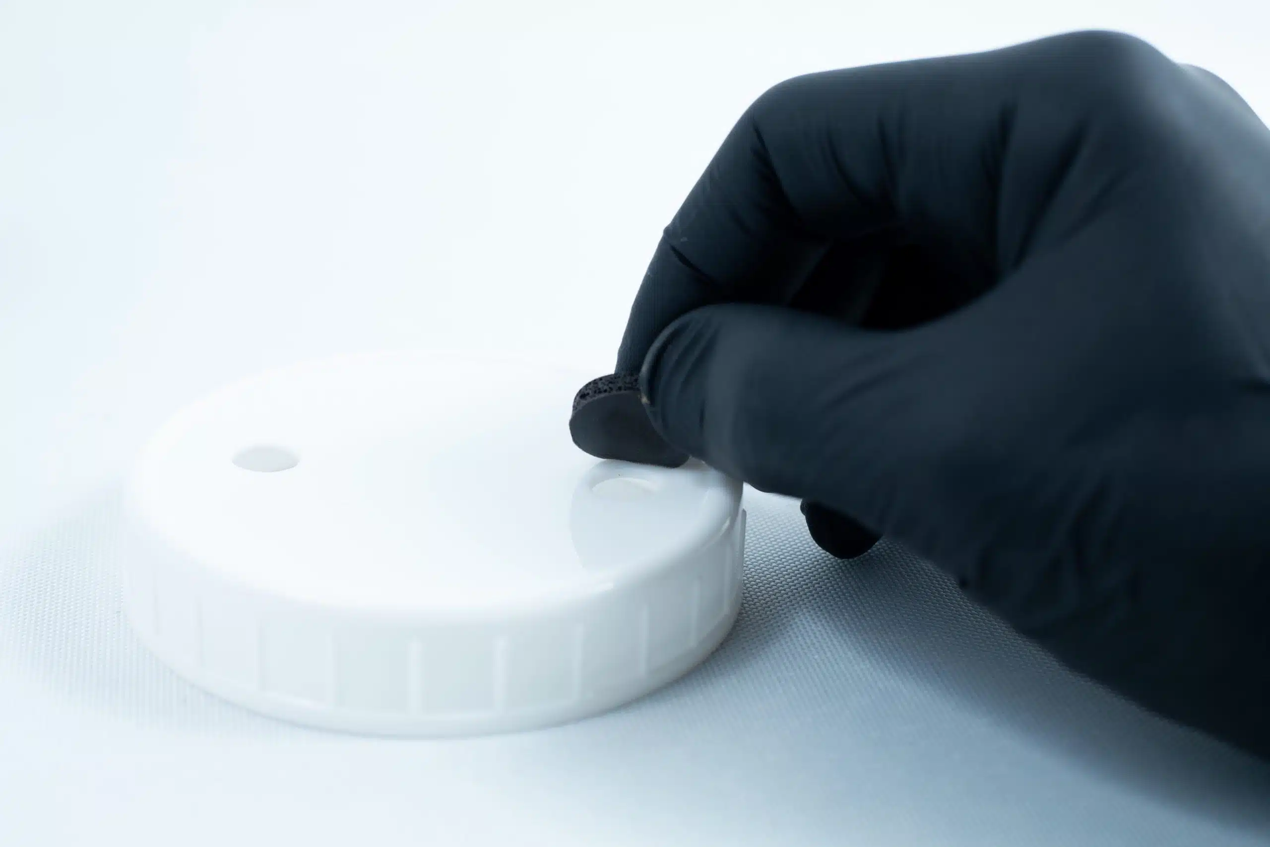 A gloved hand applies Primer 94 multisurface for 3M glue 10ML onto a round white plastic lid to prepare it for optimal tape adhesion.