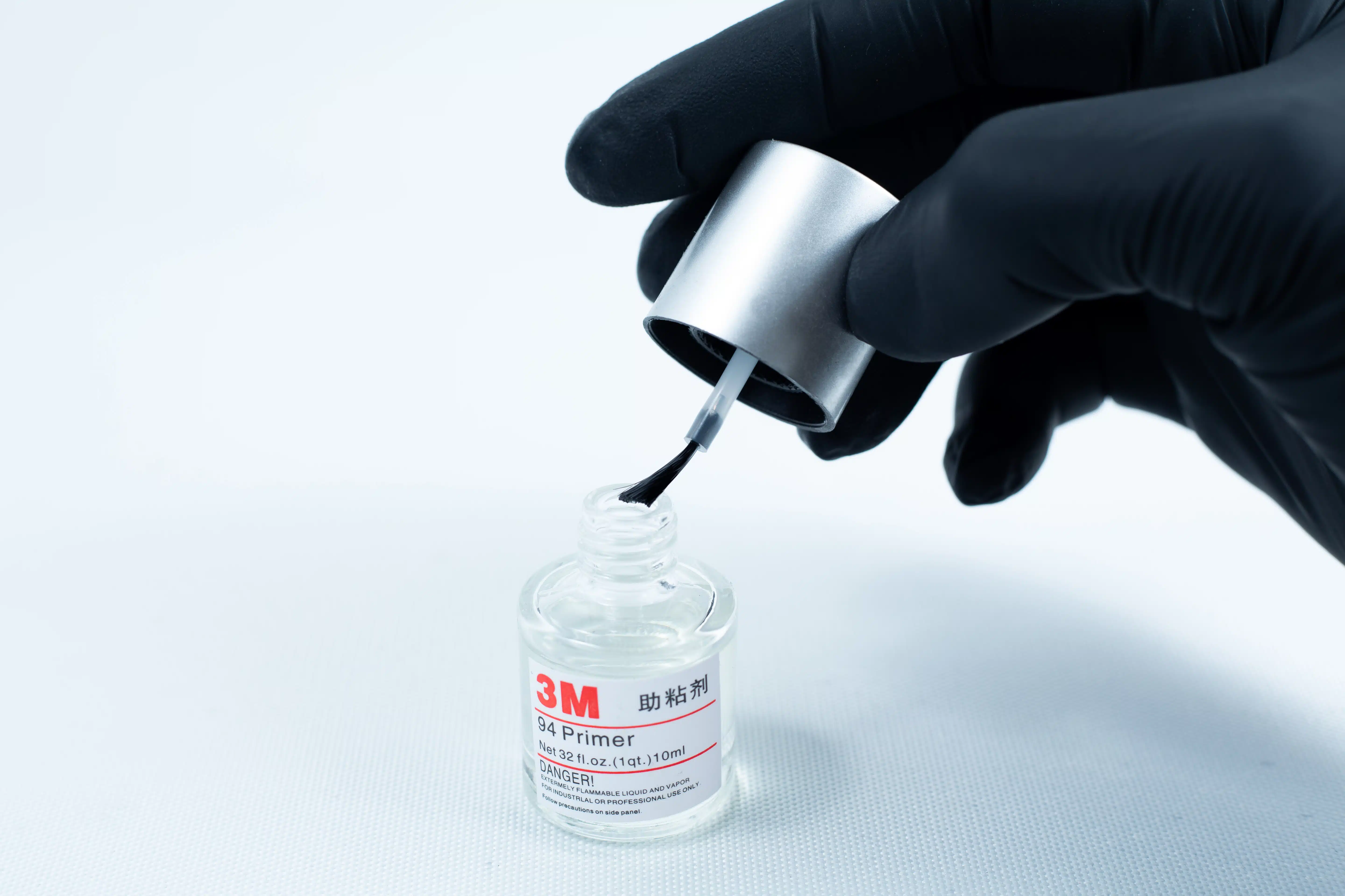 A gloved hand holds a brush applicator over an open bottle of Primer 94 multisurface for 3M glue 10ml on a white background.