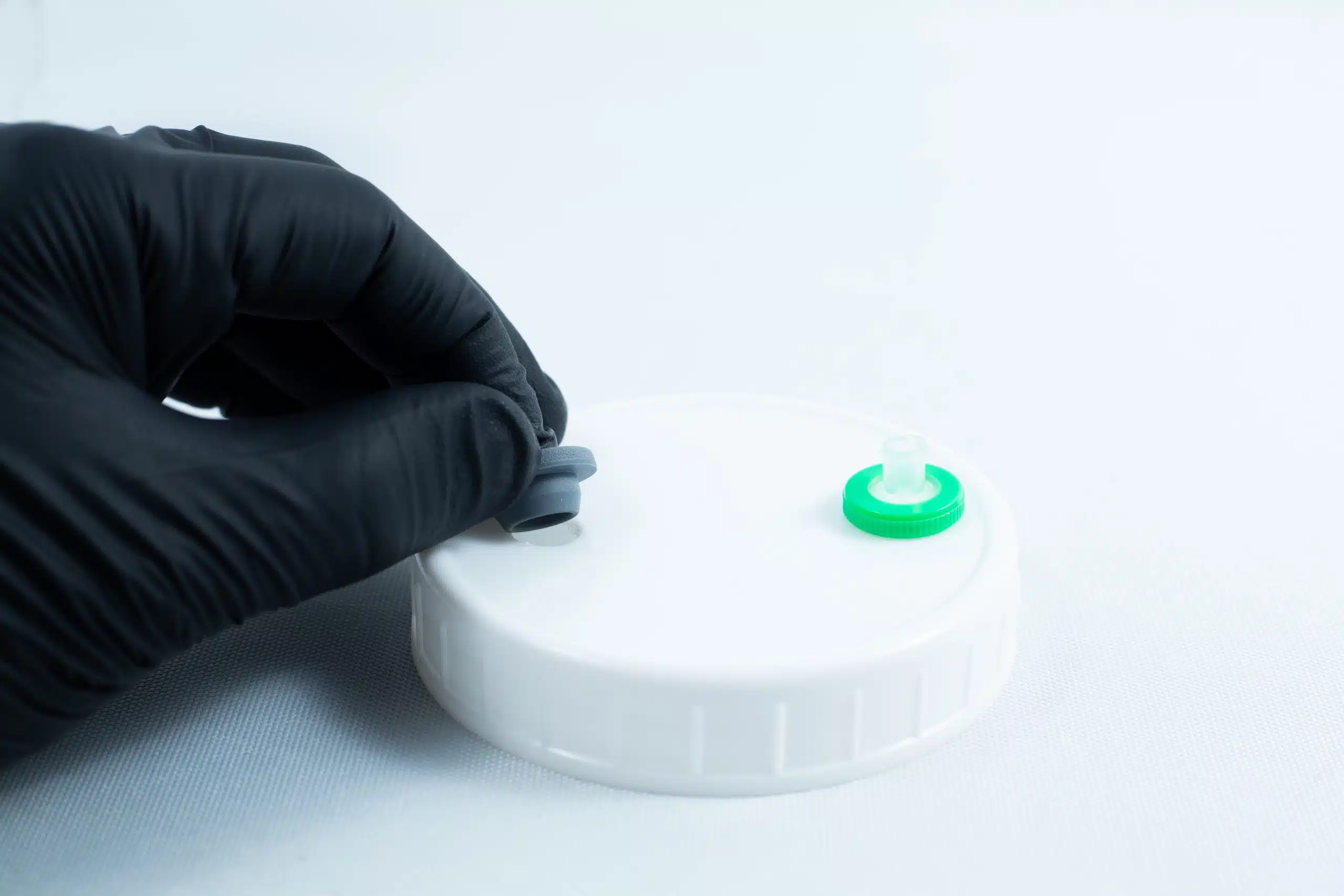 A gloved hand holds the grey cap of Primer 94 multisurface for 3M glue 10ML near a white plastic lid with a green port, commonly used as a tape primer on clean white surfaces.