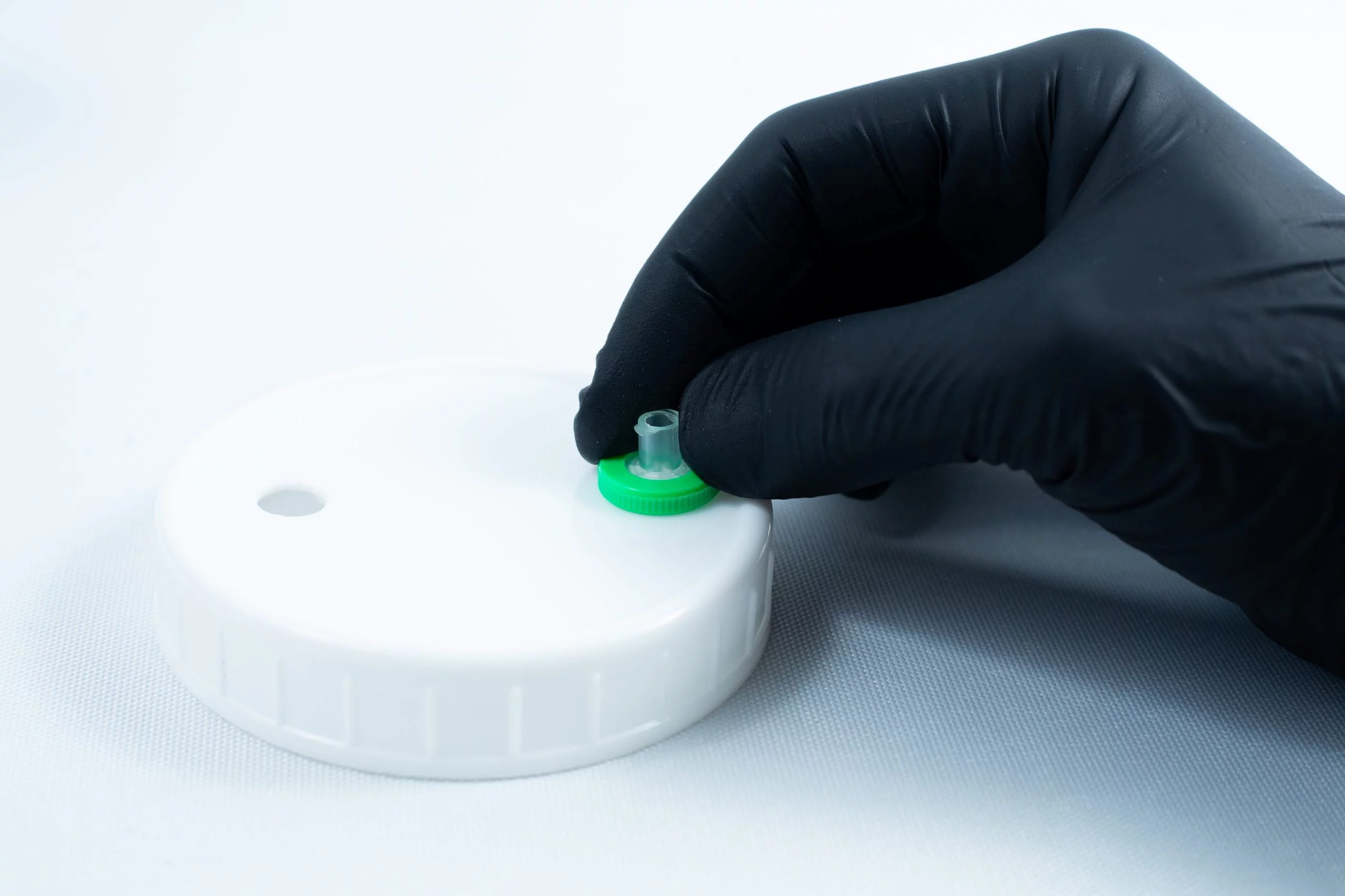 A gloved hand holds a green-capped valve on a white plastic lid, commonly used for storing Primer 94 multisurface for 3M glue 10ml, against a white background.