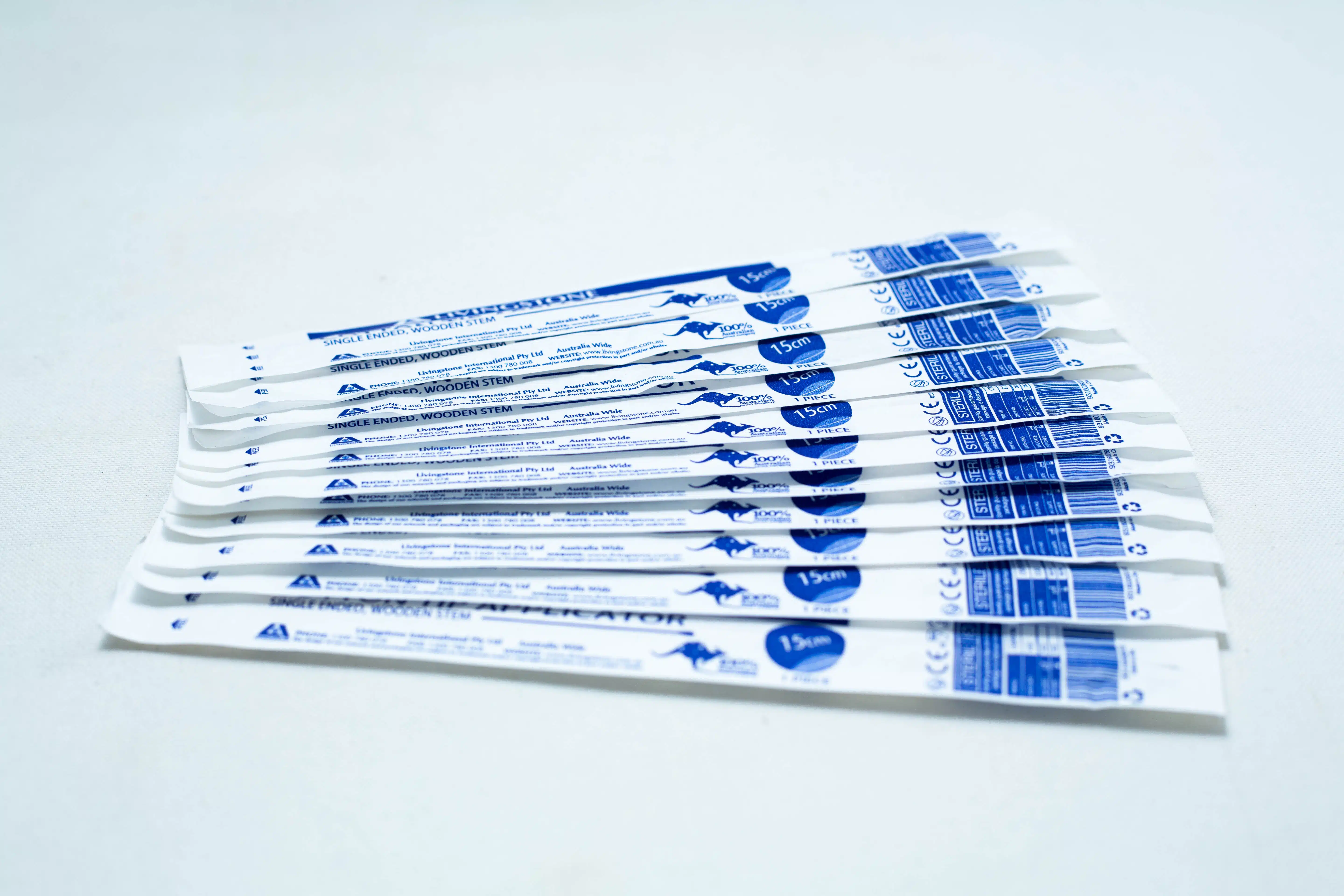Several individually wrapped sterile cotton bud products in white and blue packaging are arranged on a white surface.