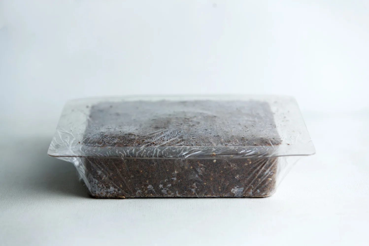 A rectangular block of soil in a plastic container covered with clear plastic film.