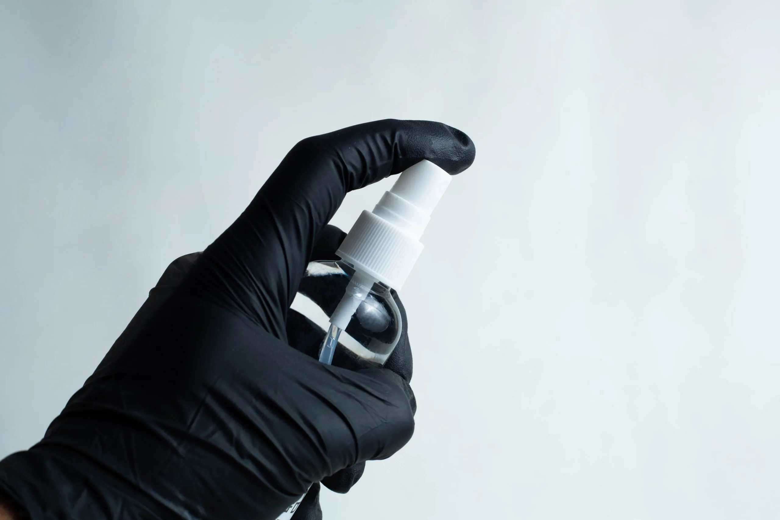 A hand in a black glove holds a 100ml spray bottle of 70% isopropyl alcohol against a plain, light background.