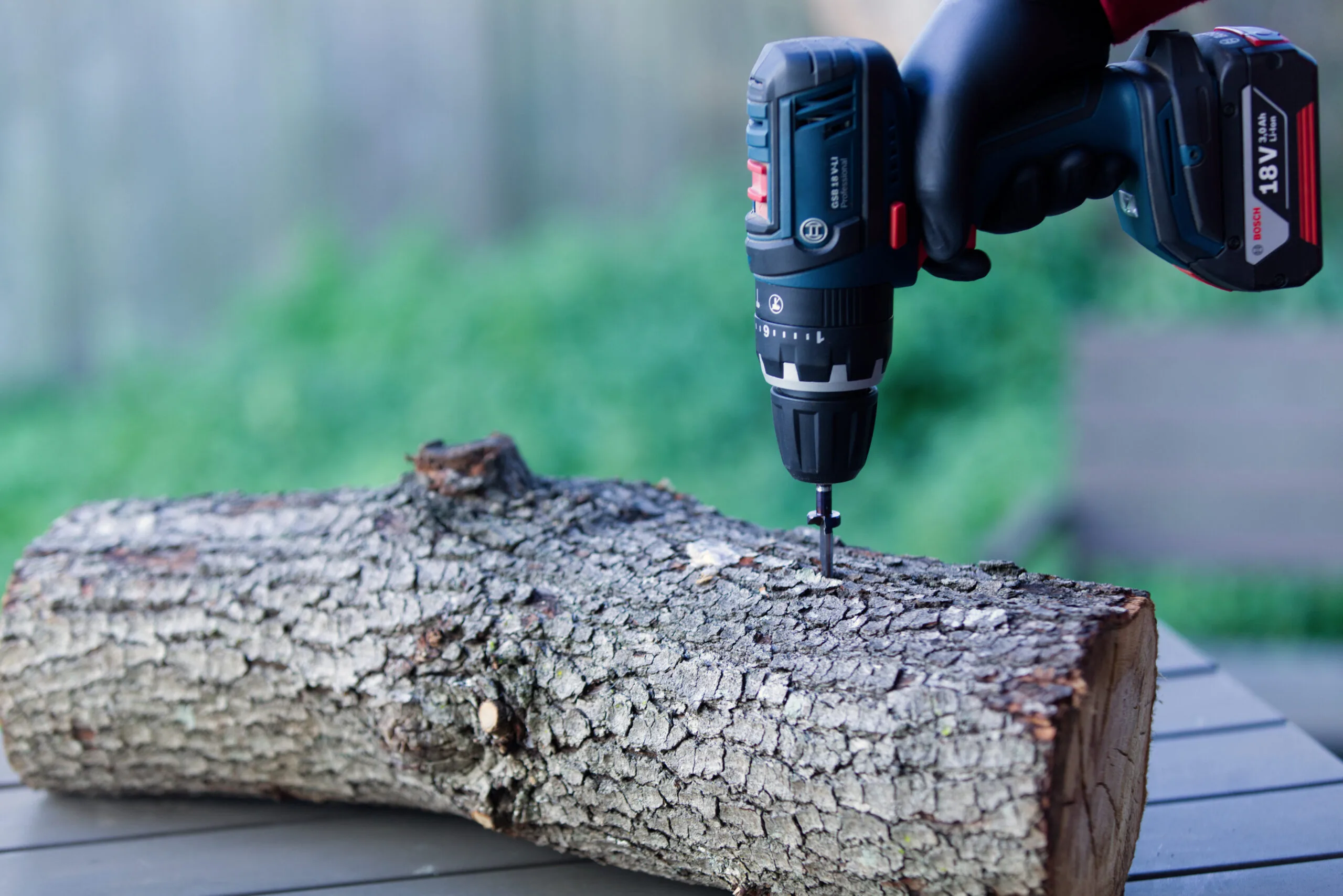 A gloved hand uses a power drill to drive a screw into a log on an outdoor table.