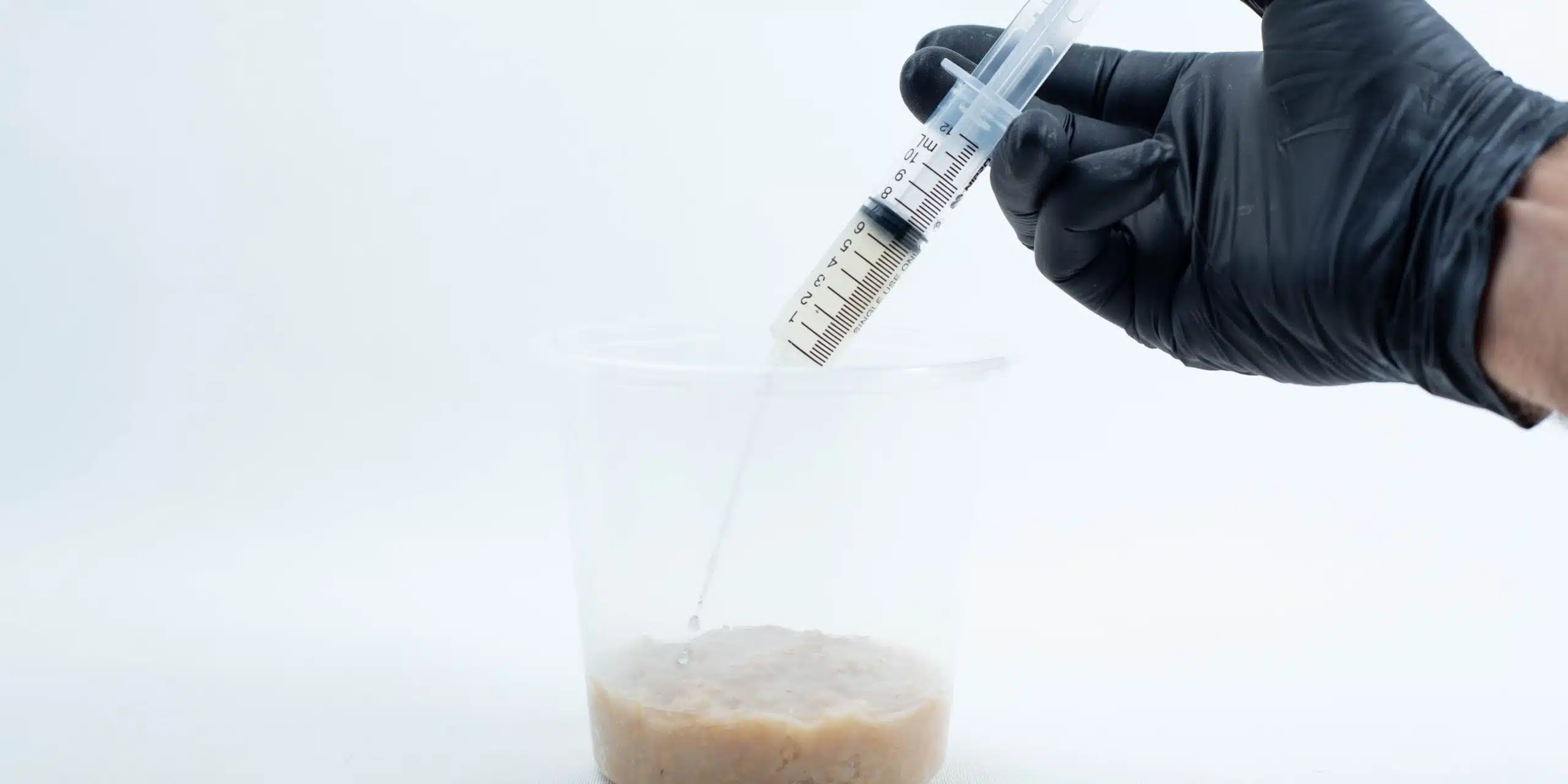 A gloved hand holds a syringe above a plastic container with a beige, lumpy liquid inside.