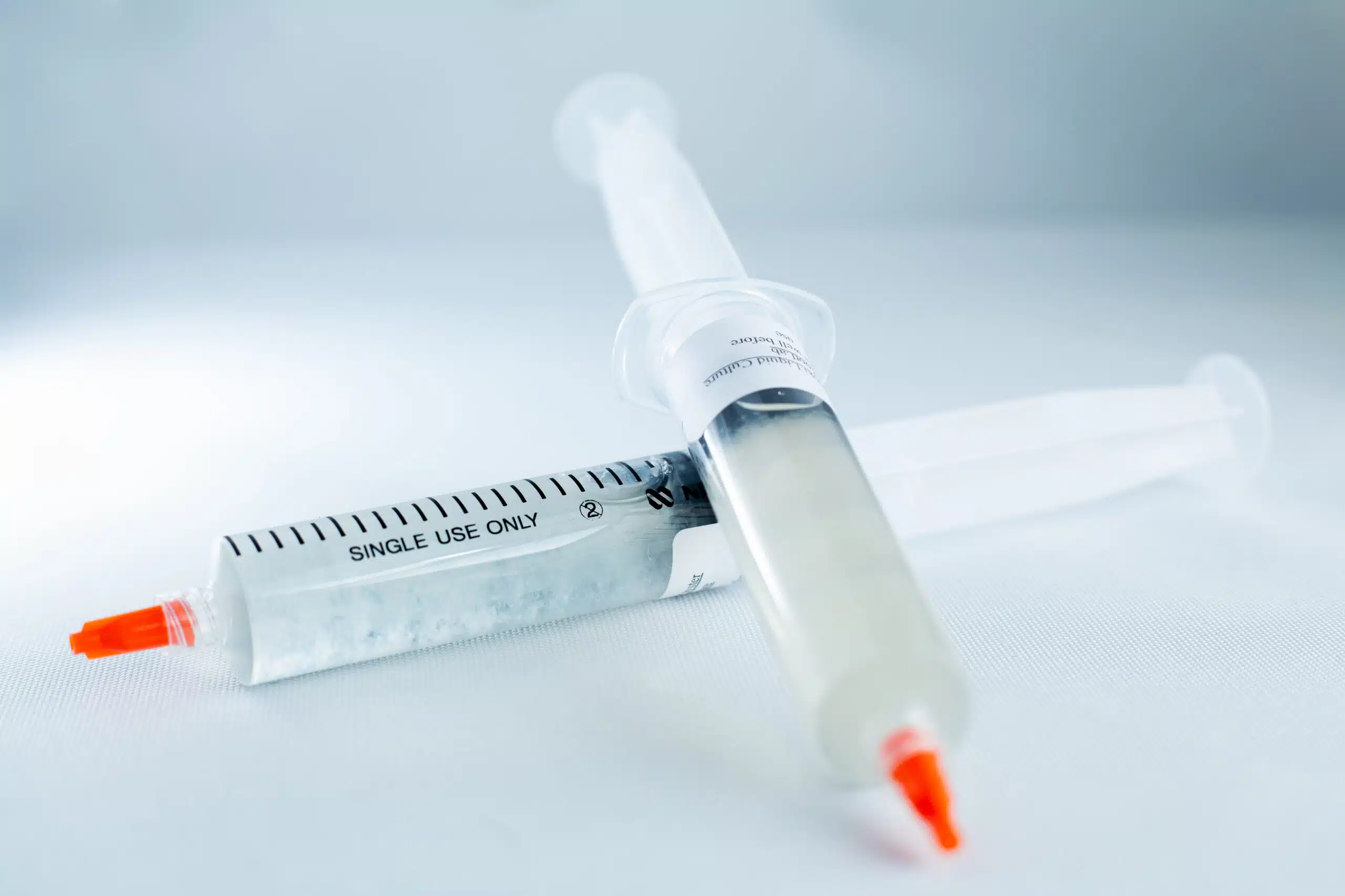 Two single-use syringes with orange caps containing Mushroom Liquid Culture | Wood Ear | Auricularia auricula-judae lie on a white surface, one filled with a clear liquid.