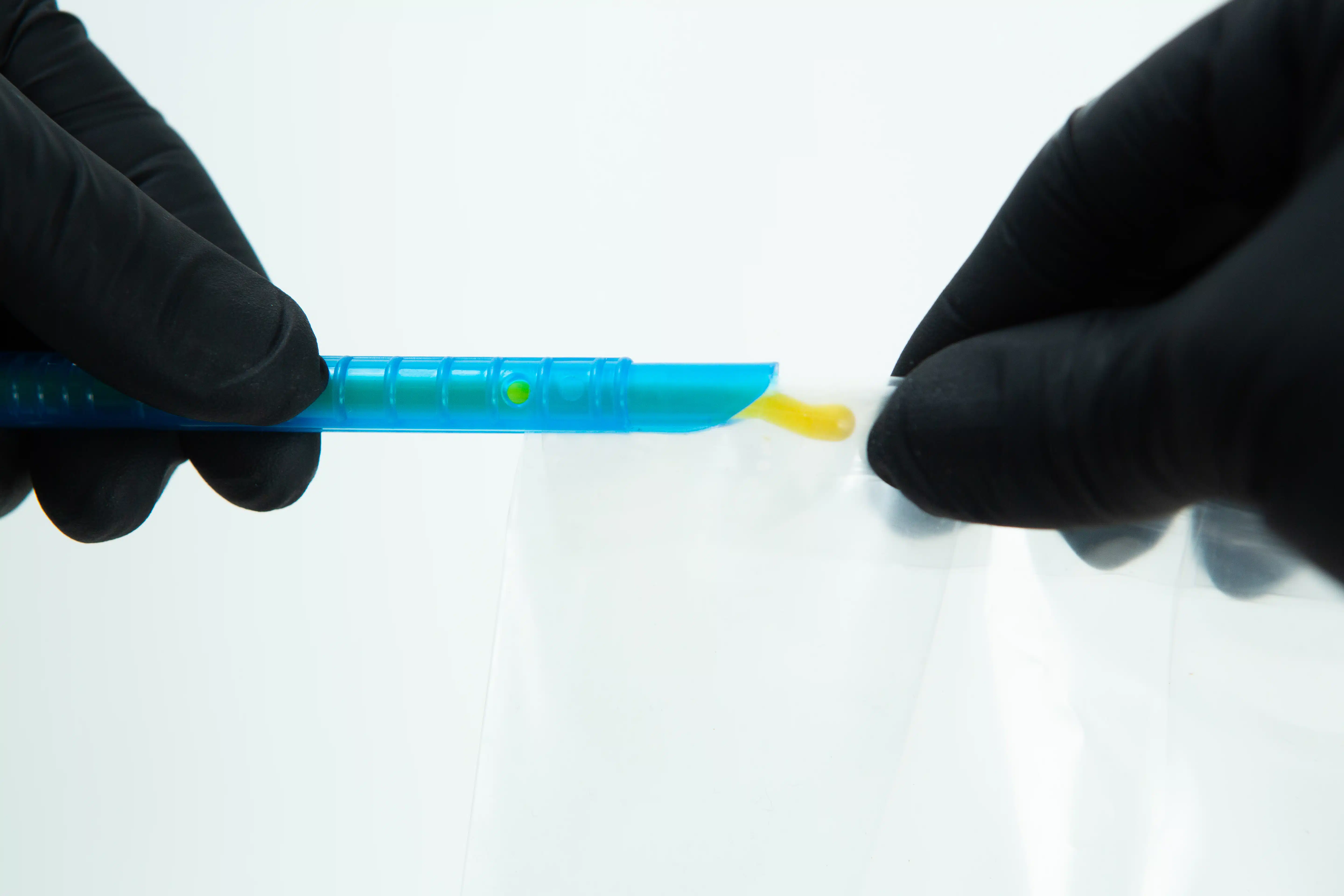Gloved hands handling a blue plastic tool with yellow substance over a clear plastic bag.
