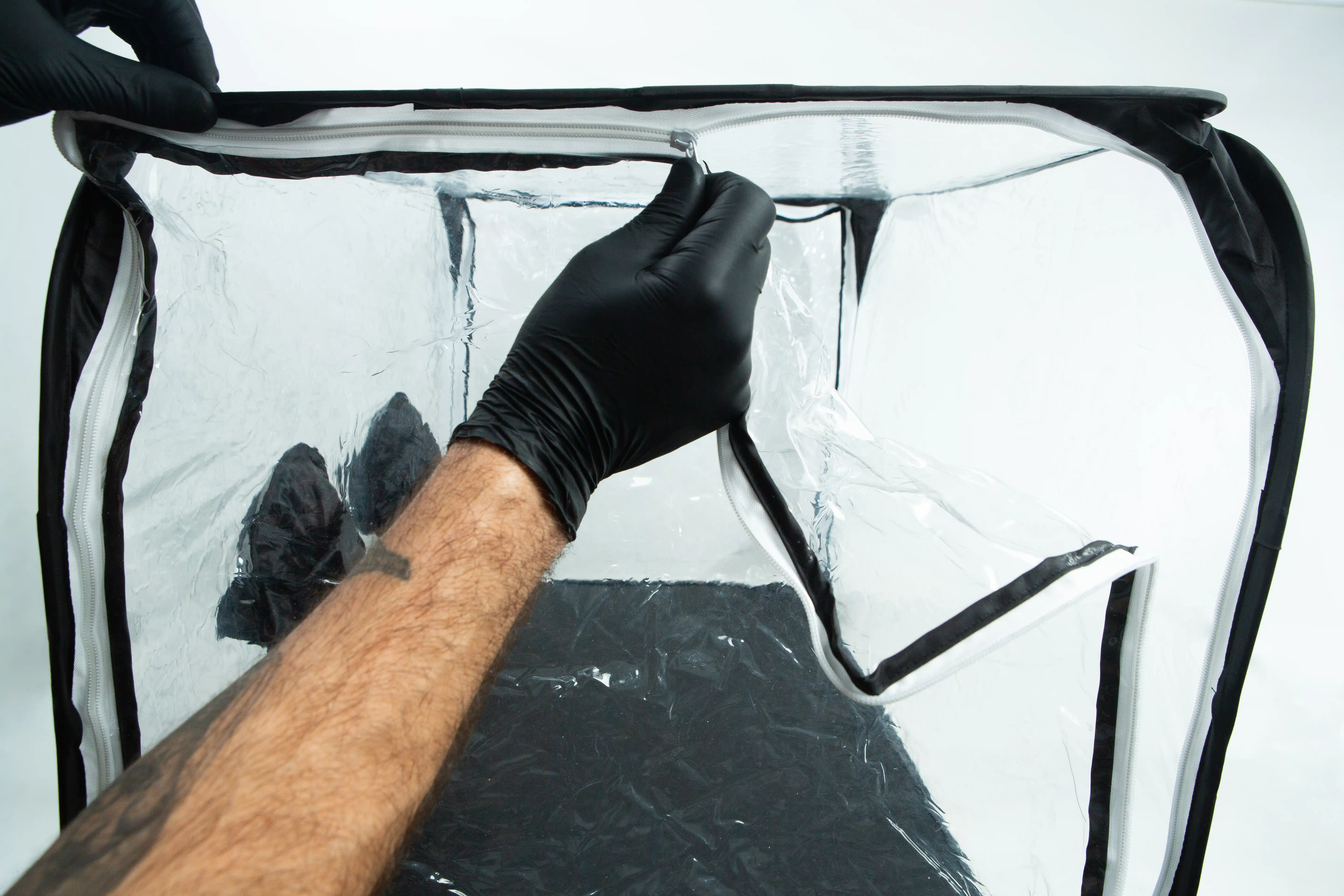 A person in black gloves unzipping a clear plastic enclosure with a black frame.
