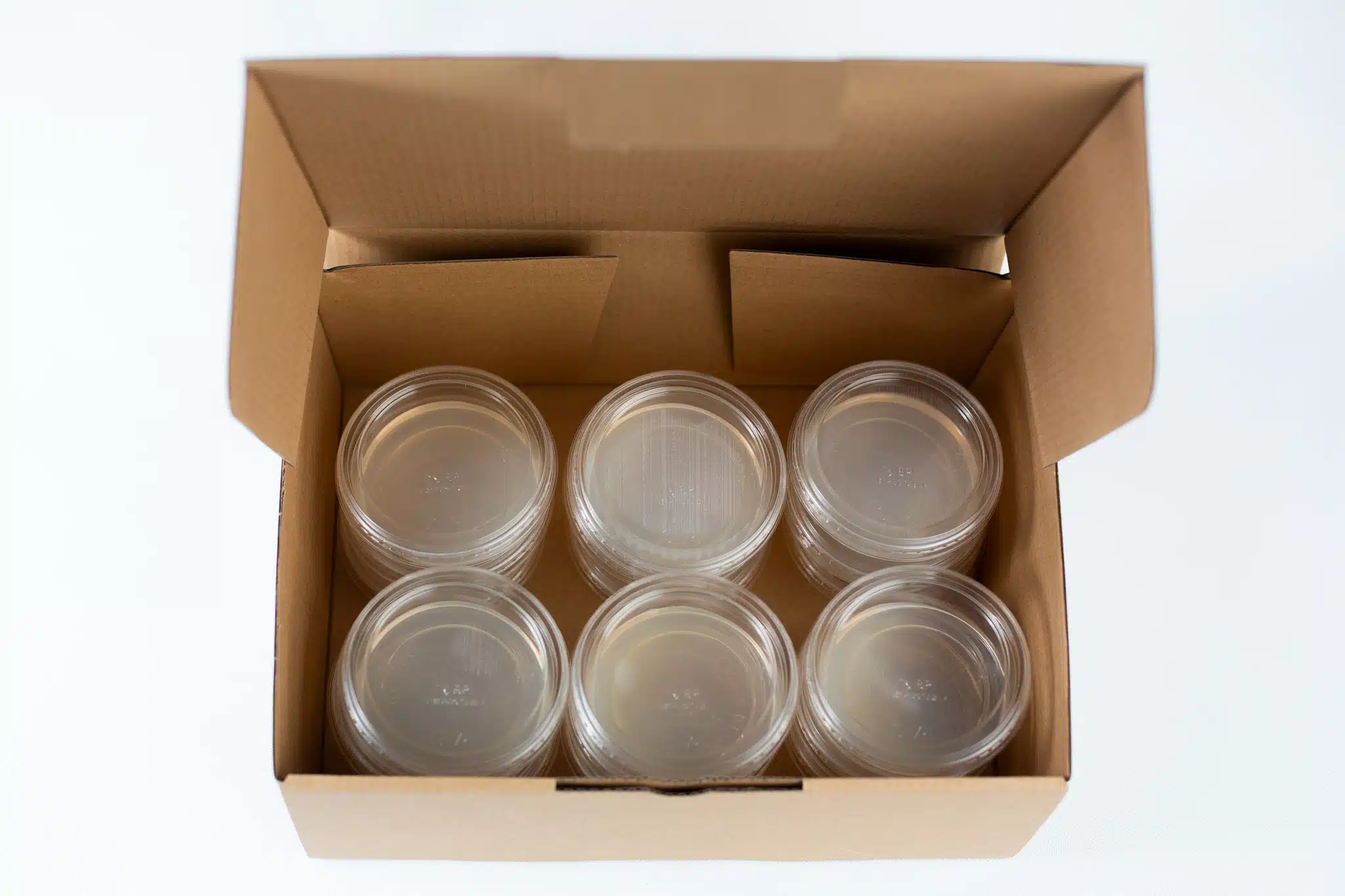 Six clear plastic petri dishes with lids in a brown cardboard box, viewed from above.
