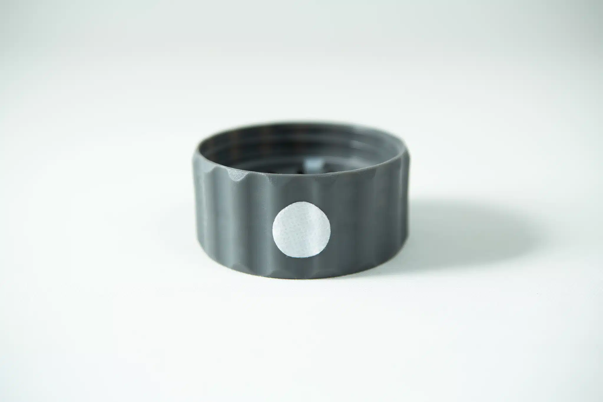 A Plant Cell Biocoupler™ Glass Set bottle cap—grey plastic with a white circular sticker on its side—rests on a white surface.