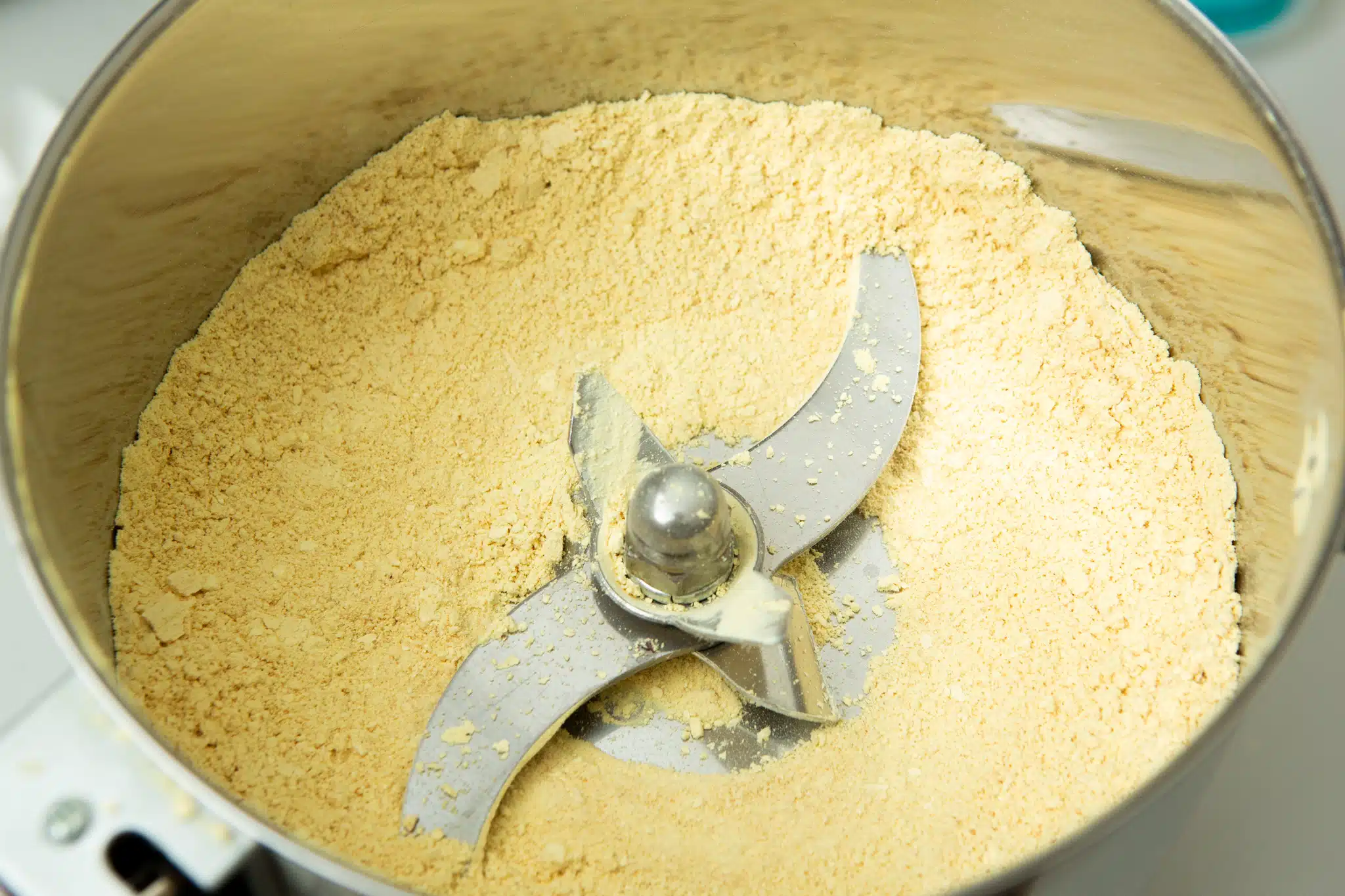 Close-up of Freeze-Dried Mushroom Lion’s Mane in a food processor, showing finely ground yellow powder and sharp metal blades inside.