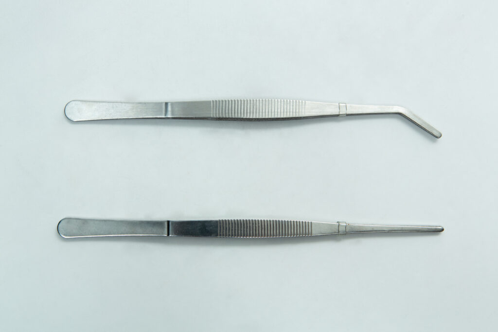 Two metal tweezers, one with a curved tip and one with a straight tip, on a plain light background.